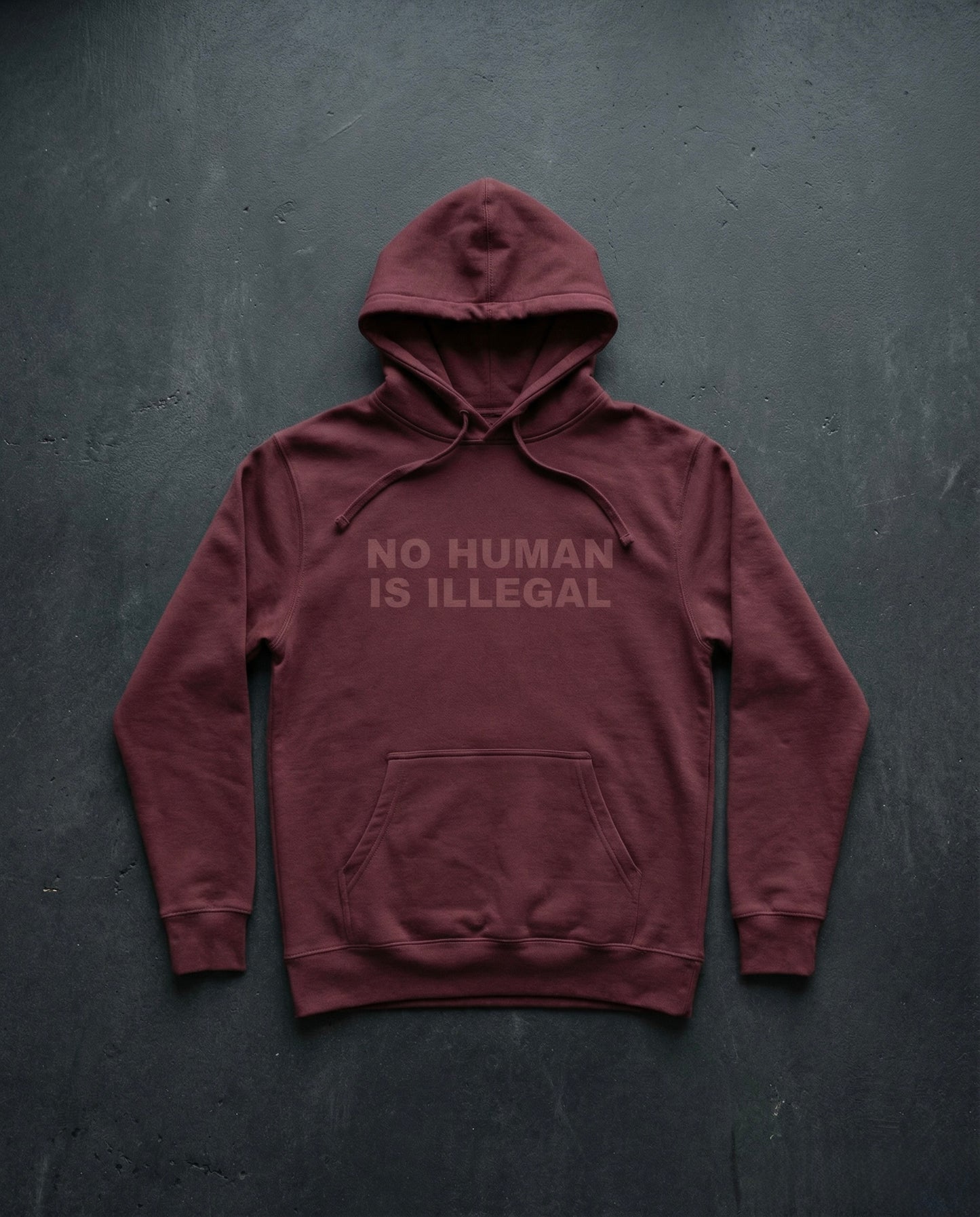 NO HUMAN IS ILLEGAL Shadow Edition Hoodie concrete background in maroon | BAD BUREAU 